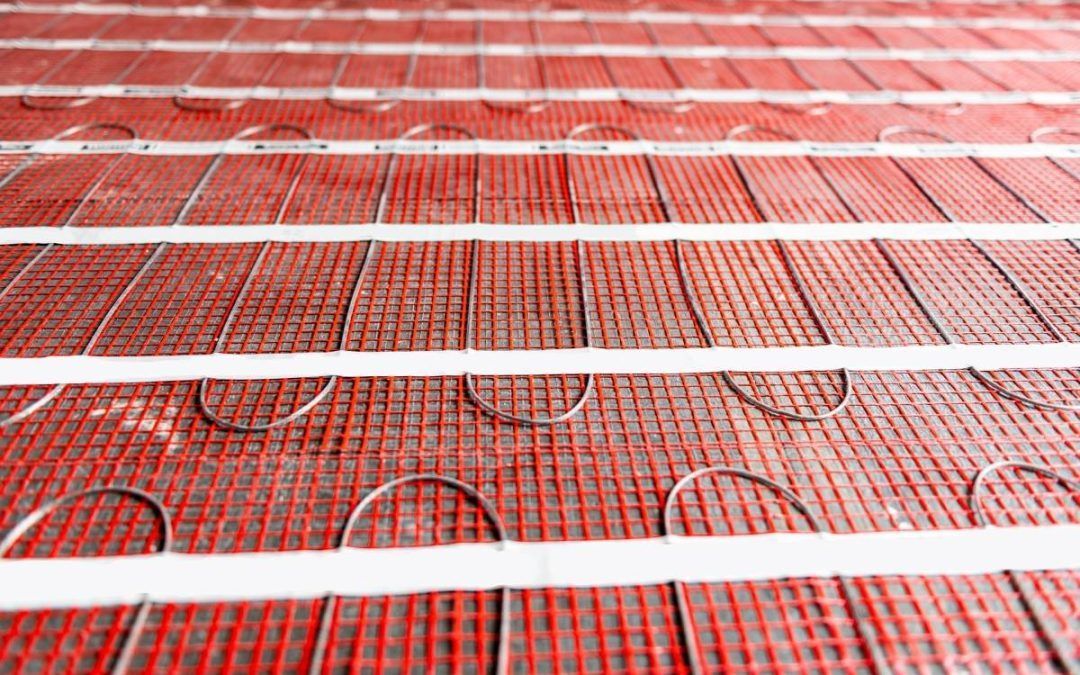 Red radiant floor heating mat with integrated electric heating wire loops secured by white adhesive strips.