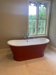 A freestanding, red, claw-foot style bathtub sits on a tiled floor in front of a window with arched detailing.