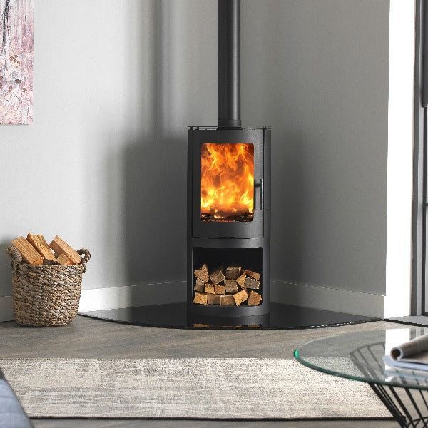 A lit black wood-burning stove sits in a corner on a glass hearth, with a basket of logs nearby in a modern room.