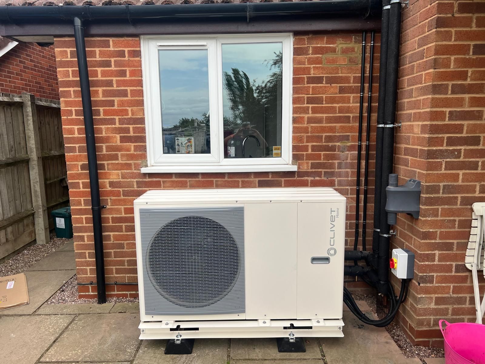 An off-white air source heat pump unit sits on a patio against a red brick exterior wall below a white window.