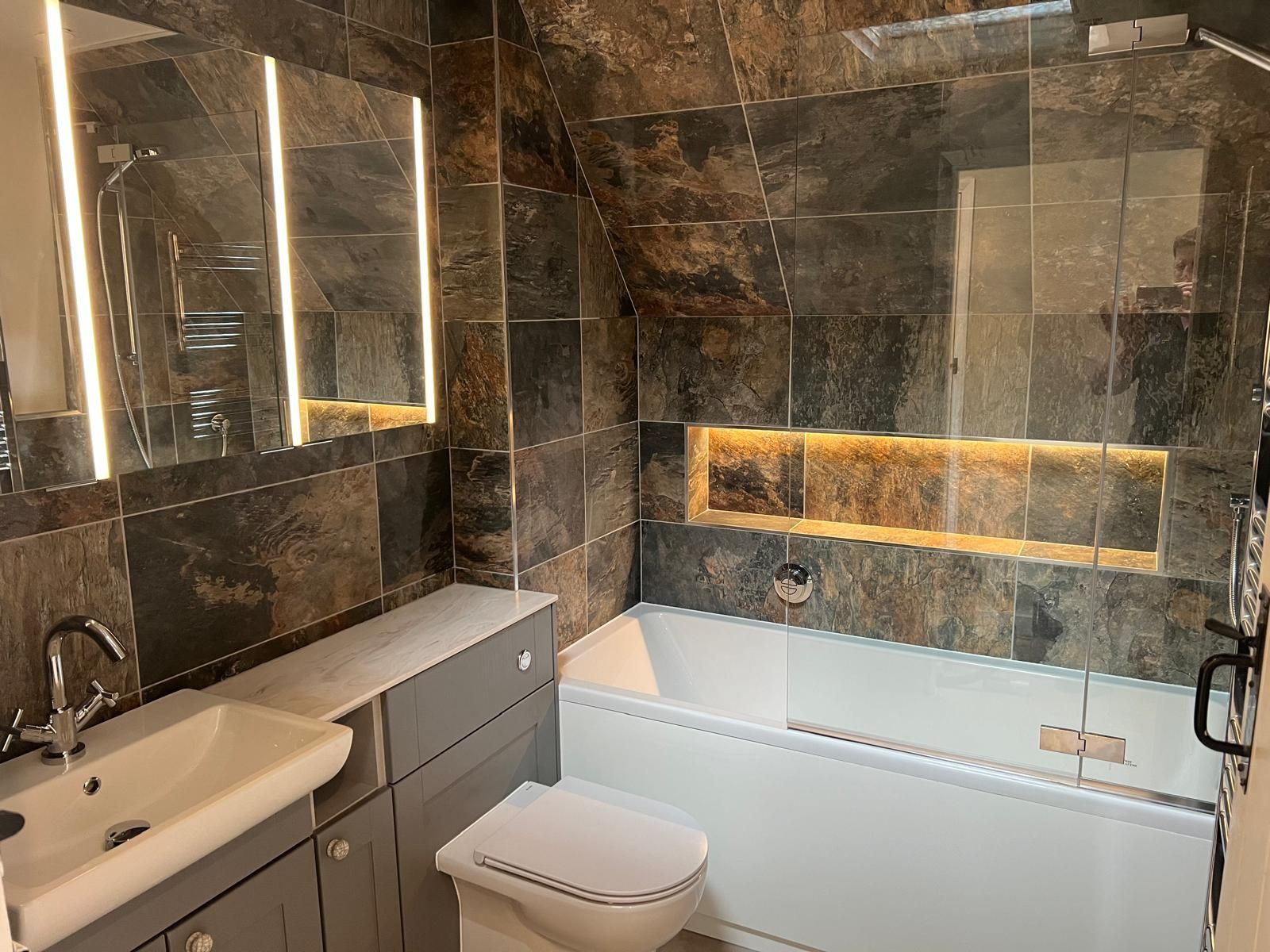 A modern bathroom with dark, textured stone tiles, a white vanity sink, a wall-mounted toilet, and a bathtub with alcove.
