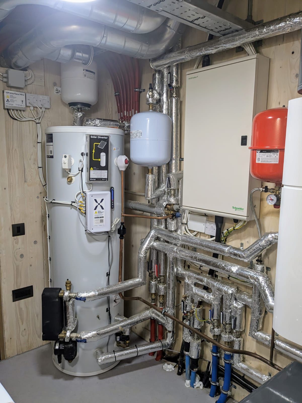 Utility room wall featuring a large water cylinder, insulated pipes, a white control panel, and red/white expansion tanks.