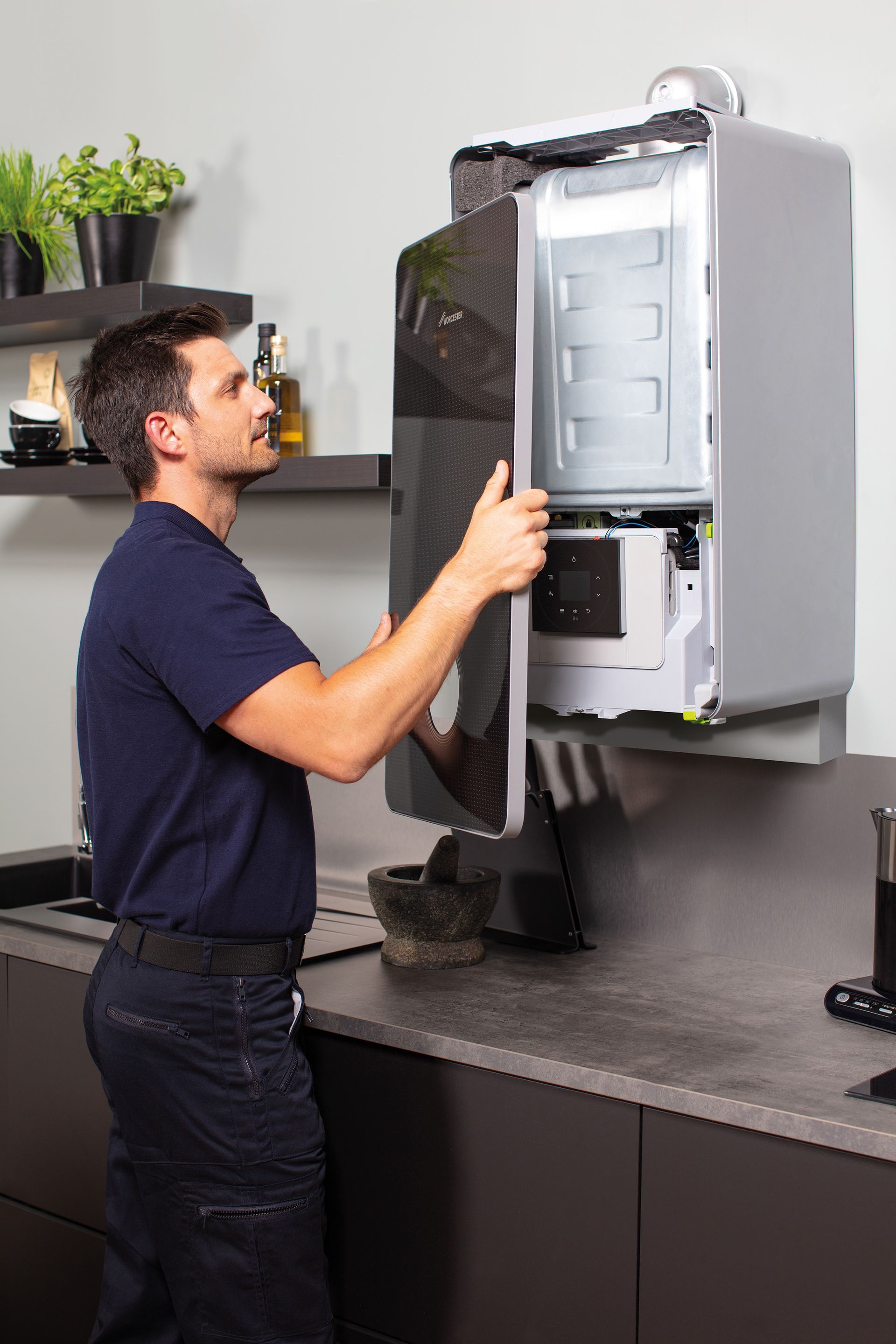 A technician in a navy uniform removes the front cover of a wall-mounted gas boiler in a kitchen.