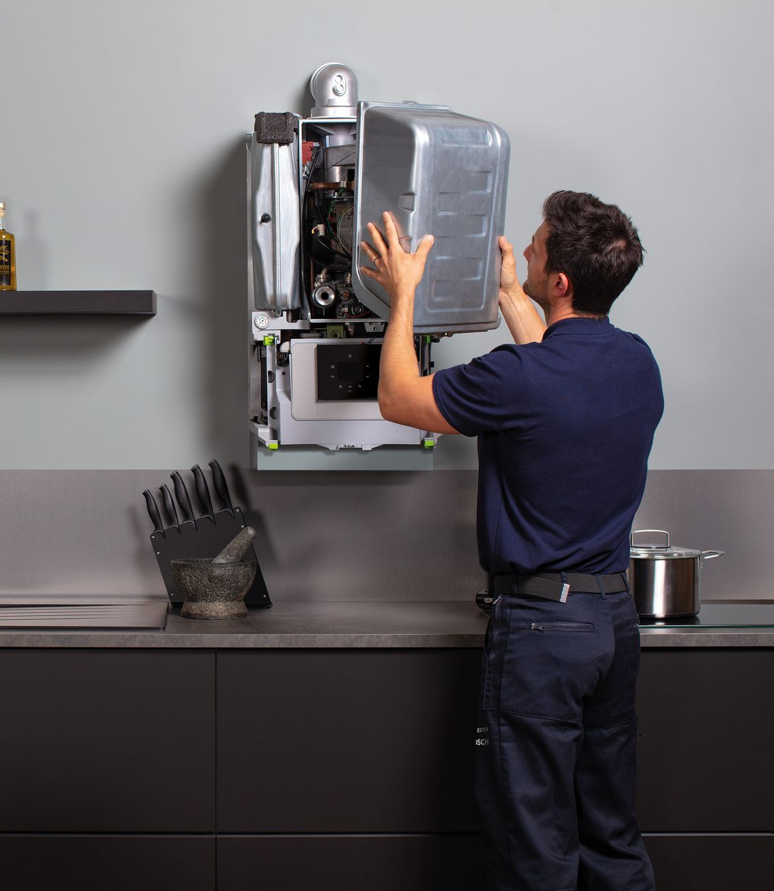 A professional wearing dark clothing attaches a metal cover to a wall-mounted boiler in a kitchen.