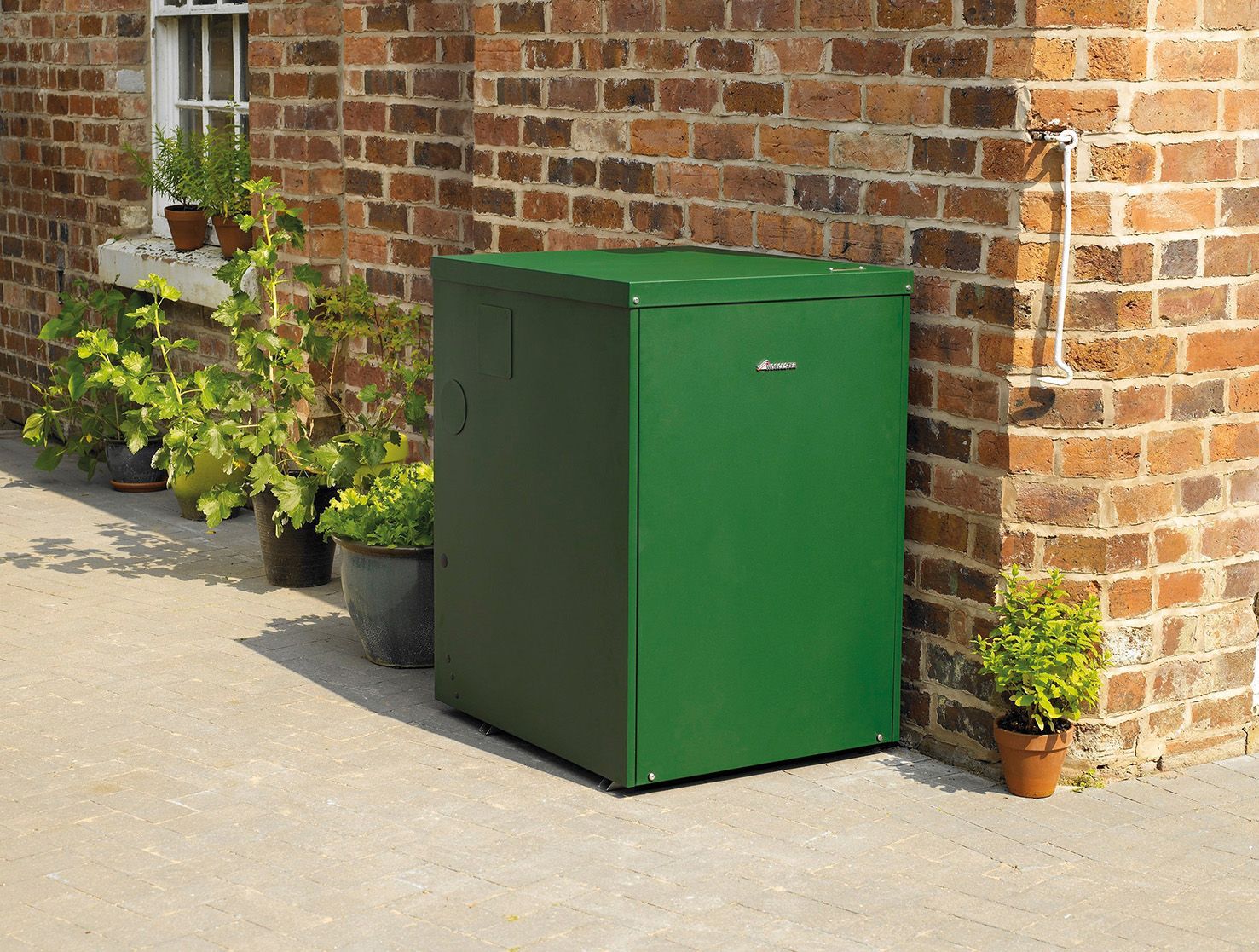 A green outdoor boiler enclosure stands against a brick wall, surrounded by several potted plants on a stone patio.