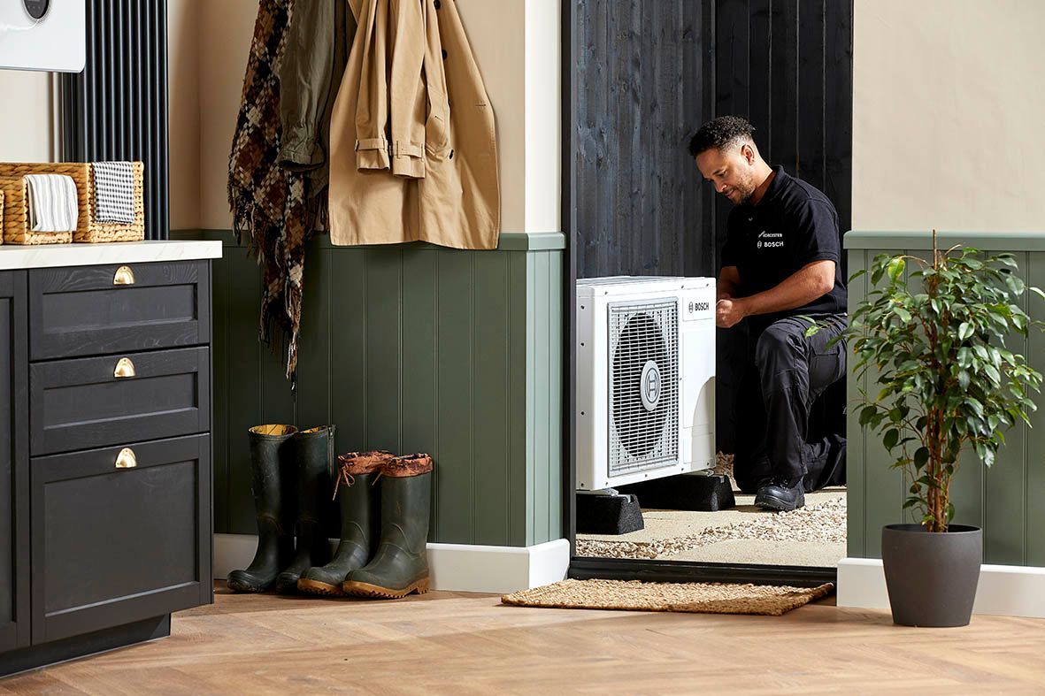 A technician kneels to install a white heat pump unit in a home interior entryway with green paneling and rubber boots.