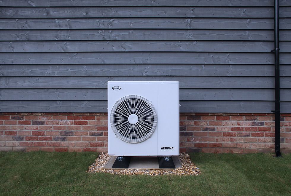 A white air source heat pump unit sits on a gravel base against a house wall with grey horizontal siding and a brick base.