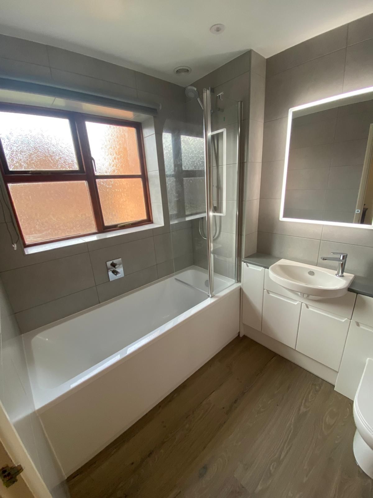 Modern bathroom with a white bathtub, glass shower screen, grey tiled walls, white vanity unit with a sink, and wood floor.