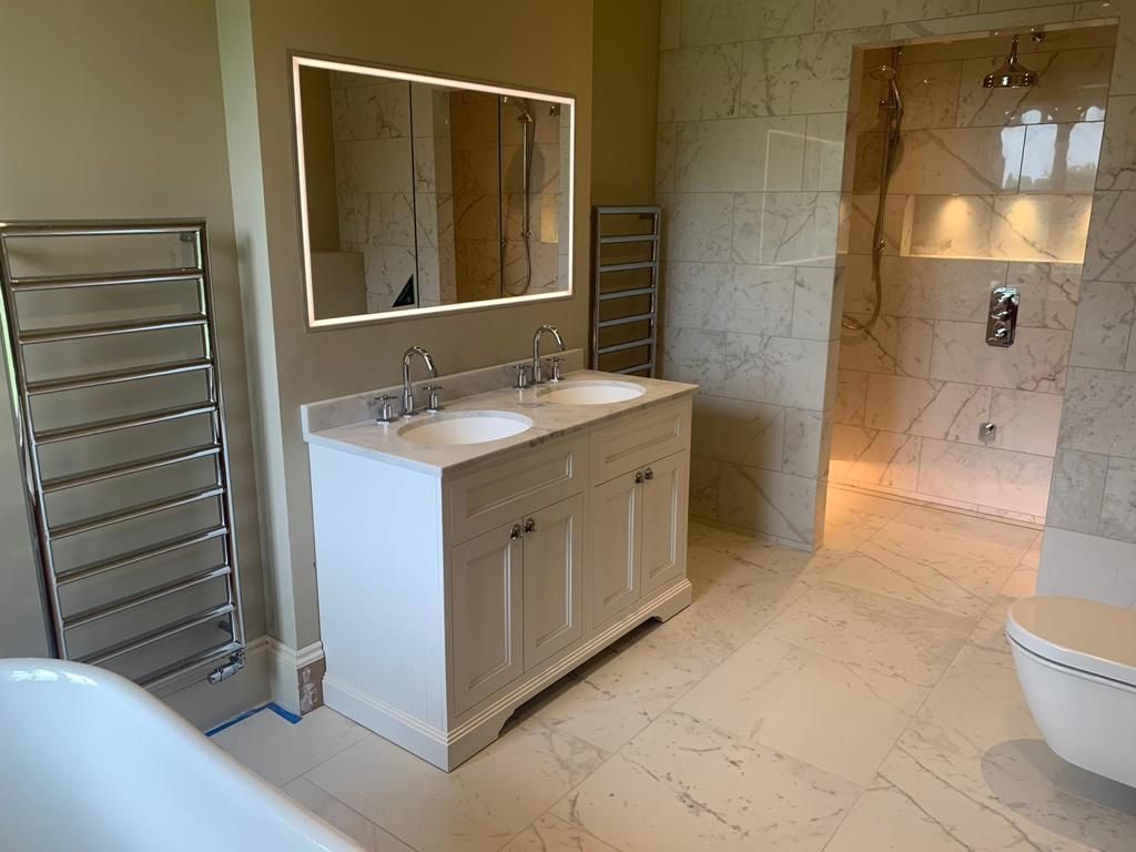 Modern bathroom featuring a double white vanity, large backlit mirror, heated towel rack, and a walk-in tiled shower.