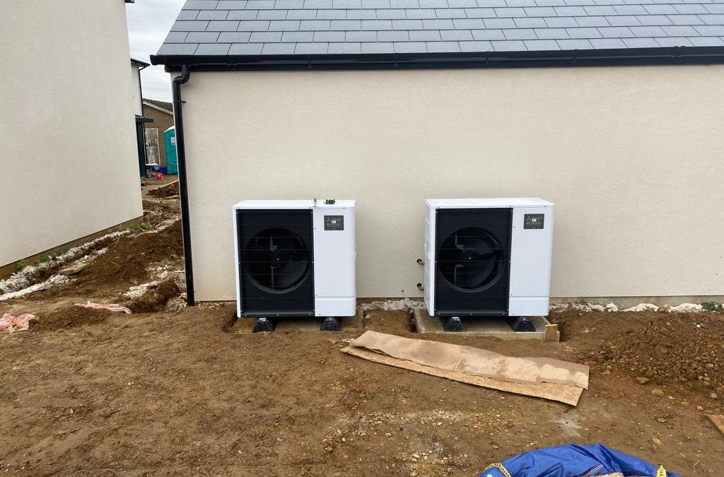 Two white air source heat pumps sit on concrete pads against the side of a light-colored building under construction.