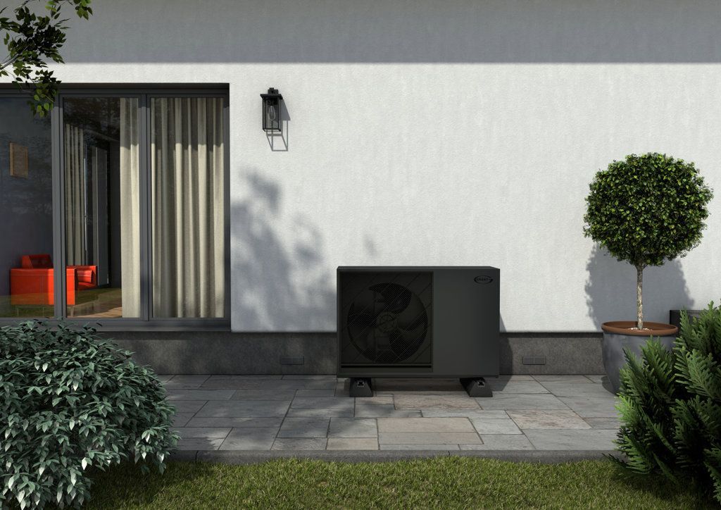 A dark gray outdoor heat pump unit sits on a stone patio against a white house wall next to a glass door and a small tree.