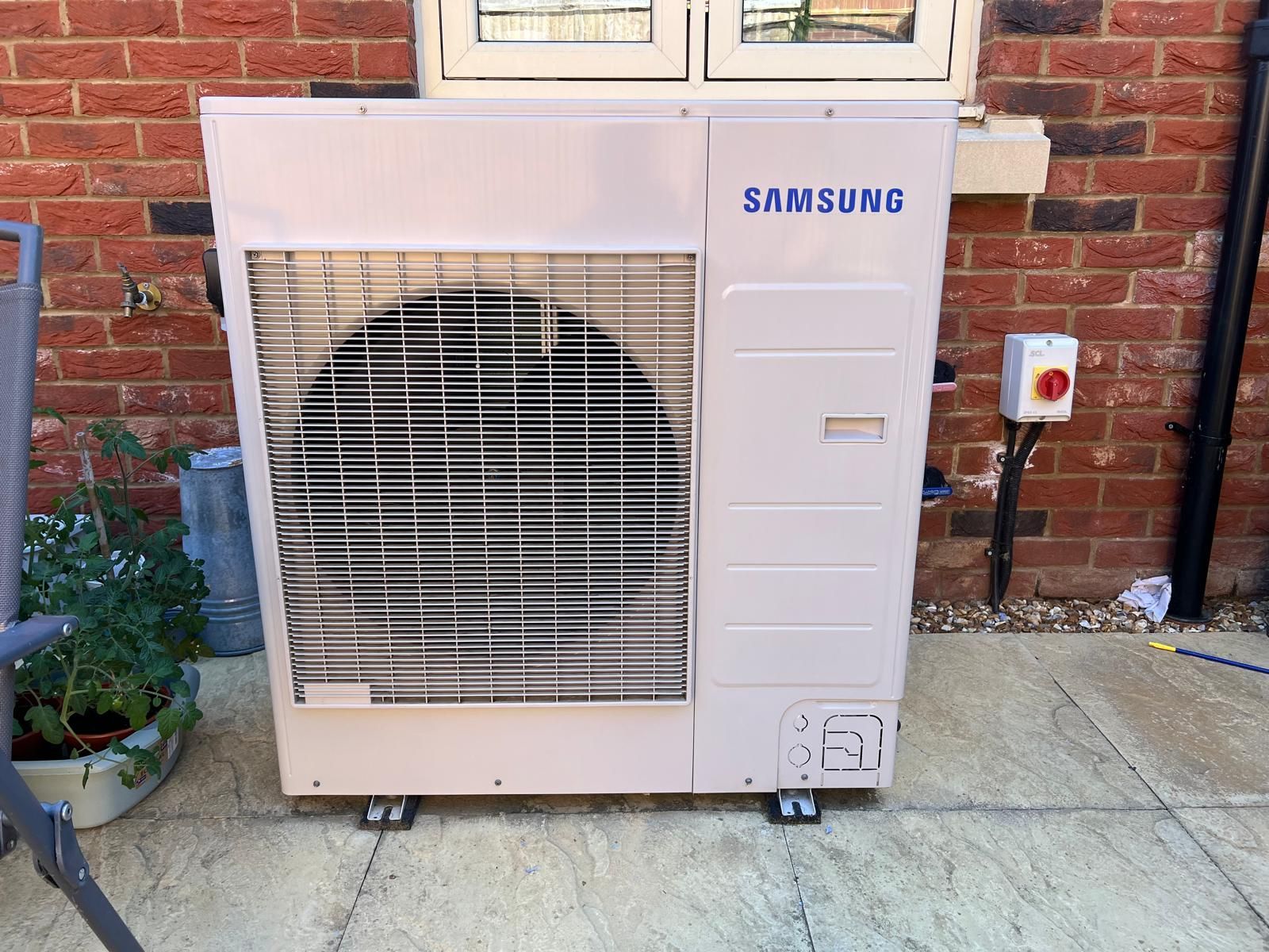 A white Samsung heat pump unit installed outdoors on a paved patio against a red brick wall.
