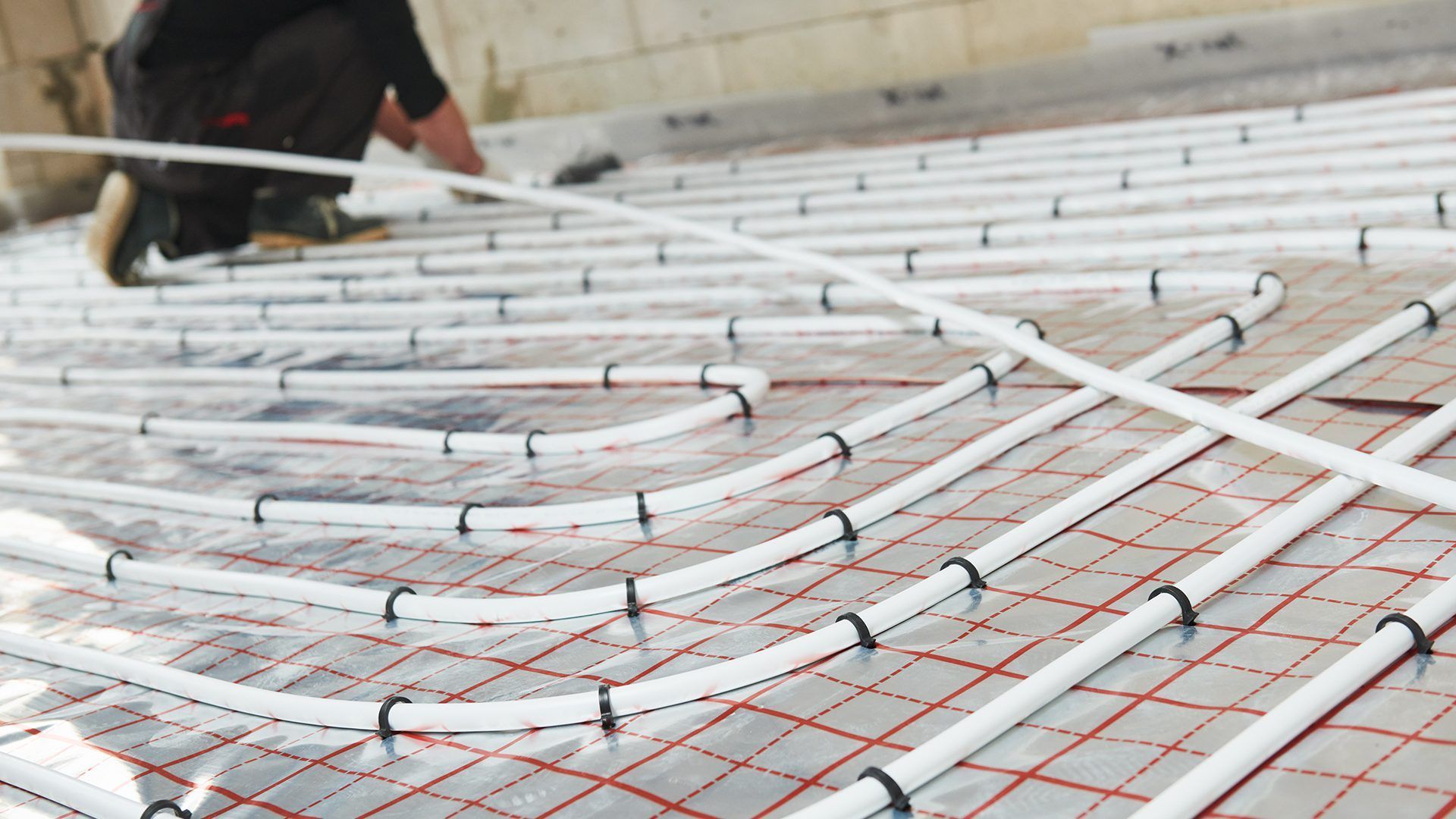 A worker installs white tubing for a radiant floor heating system on a reflective, grid-patterned underlayment.