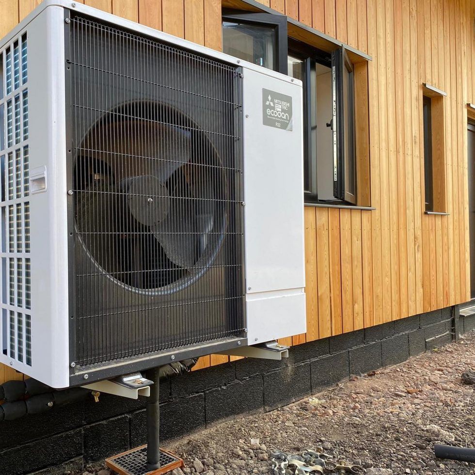 A white Mitsubishi Ecodan heat pump unit mounted outside on a stand, positioned against a light wood-clad wall.