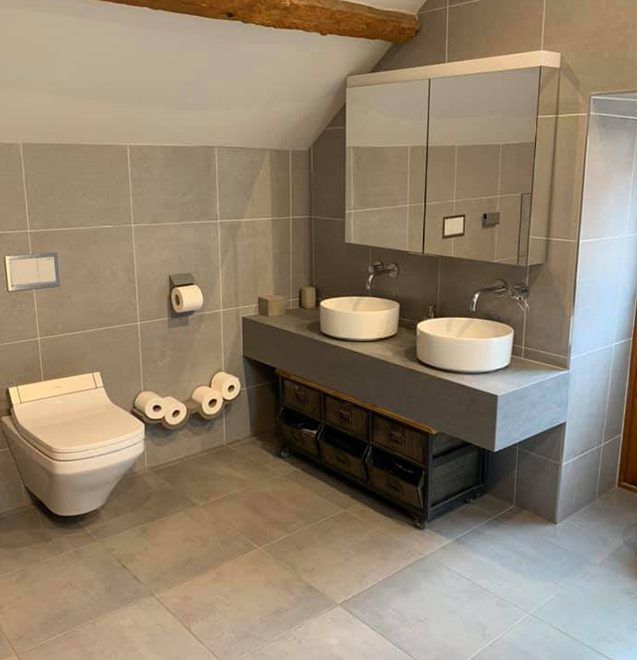 A modern bathroom with grey tiled walls, a wall-mounted toilet, and a double vanity with two white round vessel sinks.