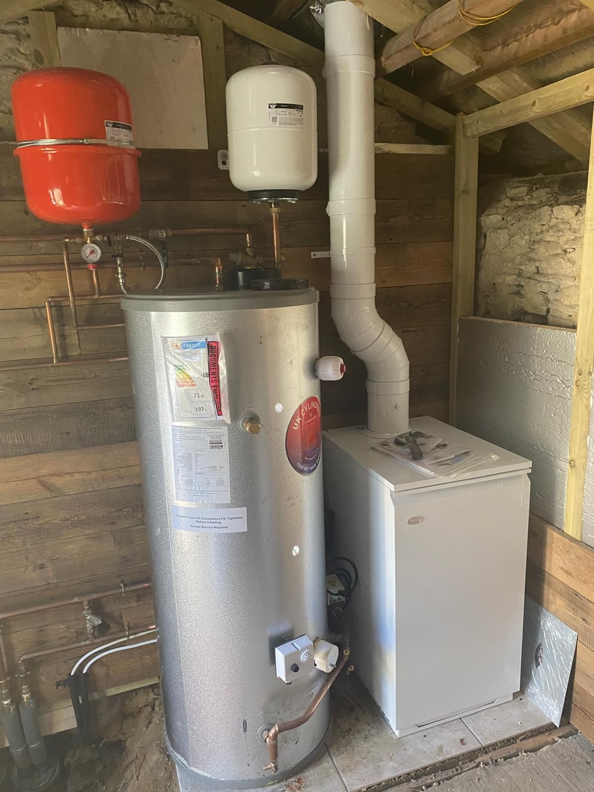 A large silver hot water cylinder, a white boiler with a flue, and two expansion vessels in an unfinished wooden room.
