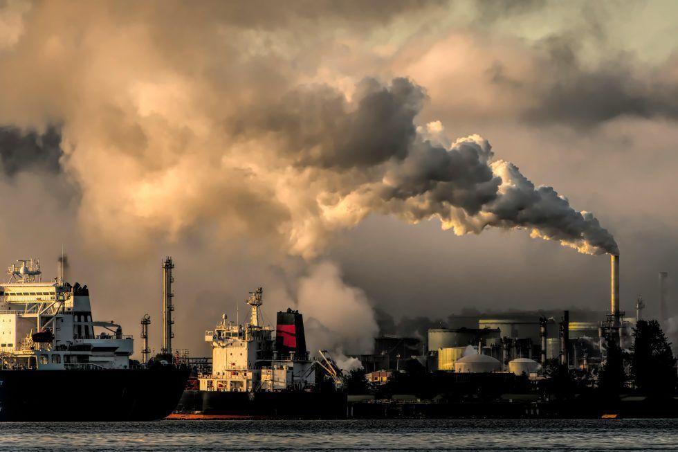 Ships dock at an industrial port facility as a tall smokestack releases thick, dark smoke into the cloudy sky.