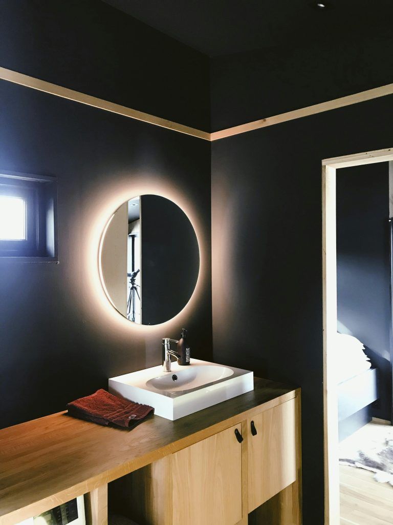 A modern bathroom with matte black walls, a light wood vanity, a square vessel sink, and a circular LED-backlit mirror.