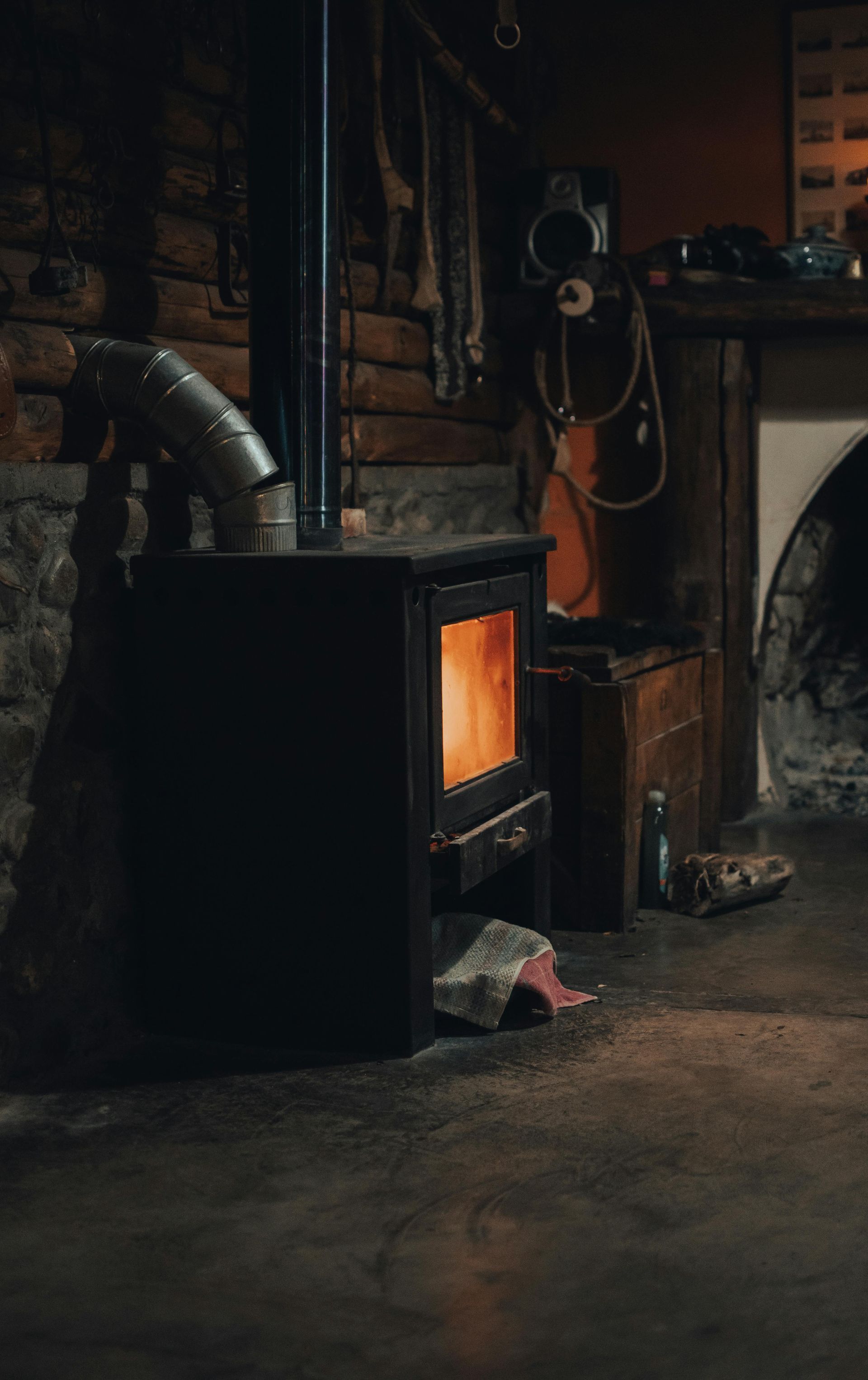 A dark, rustic room with a lit black wood-burning stove, stone walls, and hanging cords.