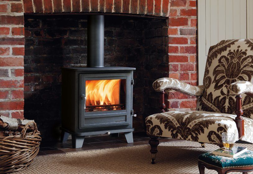 A lit wood-burning stove sits in a brick fireplace next to an ornate patterned armchair on a textured rug.