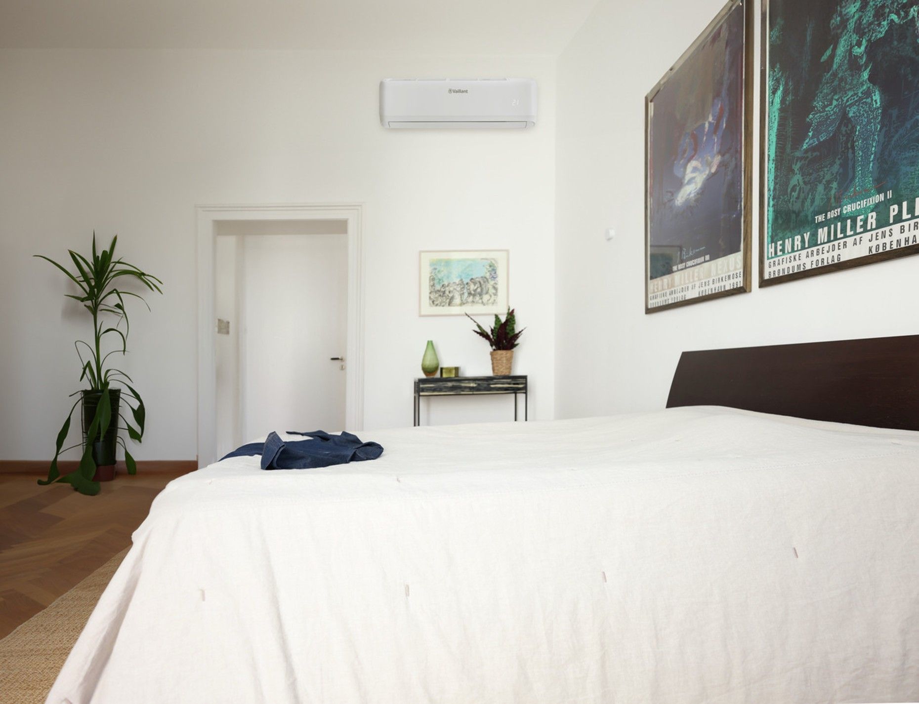 A bedroom with a white-covered bed, a tall potted plant on the left, a small side table, and two framed posters on the wall.