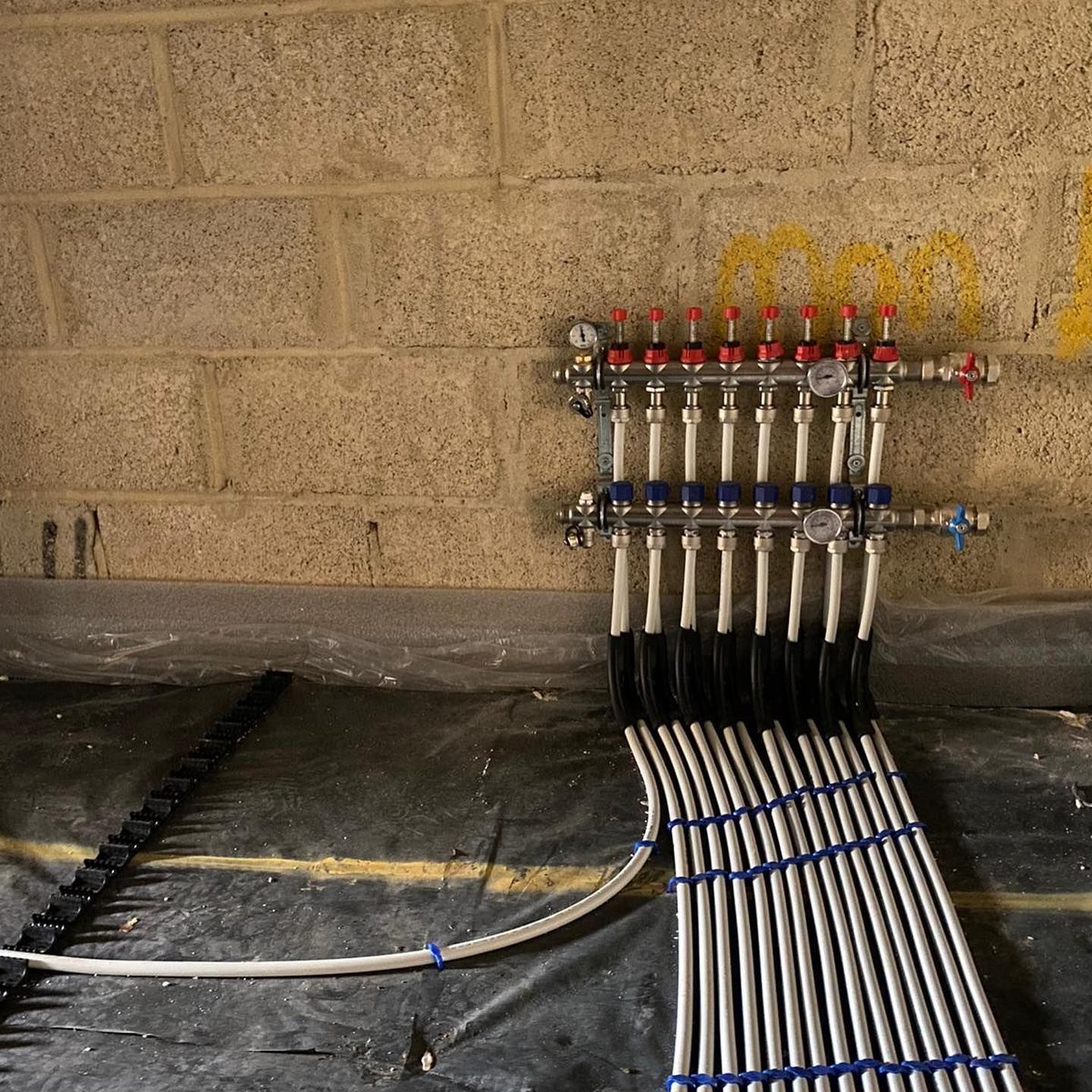 A radiant floor heating manifold with multiple pipes mounted on a cinder block wall above a floor underlayment.