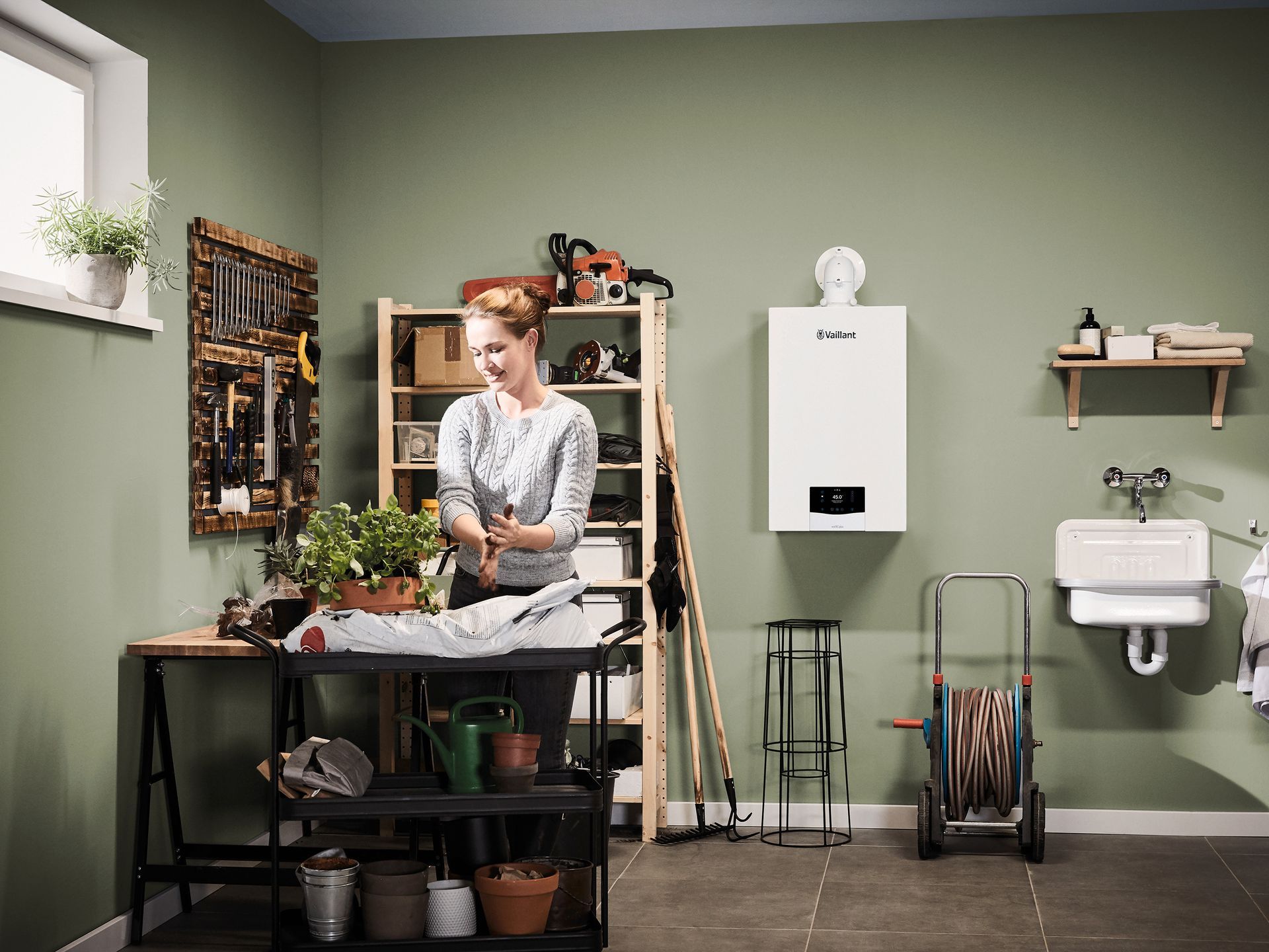 A person tends to potted plants at a workstation in a garage with green walls, shelving, a boiler, and a garden hose reel.