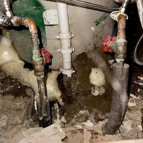Pipes with valves and insulation, covered in residue, in a cluttered, damp setting.