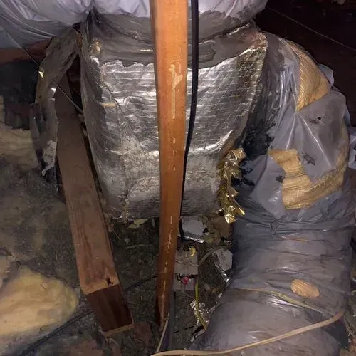 Damaged HVAC ductwork in an attic, with torn insulation and exposed metal. Wooden beams are visible.