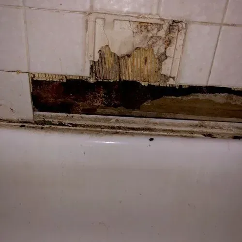 Damaged bathroom tiles above a bathtub reveal rotted wood underneath.