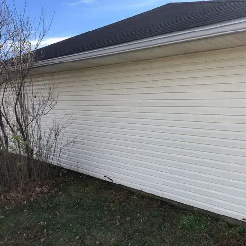 White vinyl siding on a building's side, dark roof, and sparse bush against a blue sky.