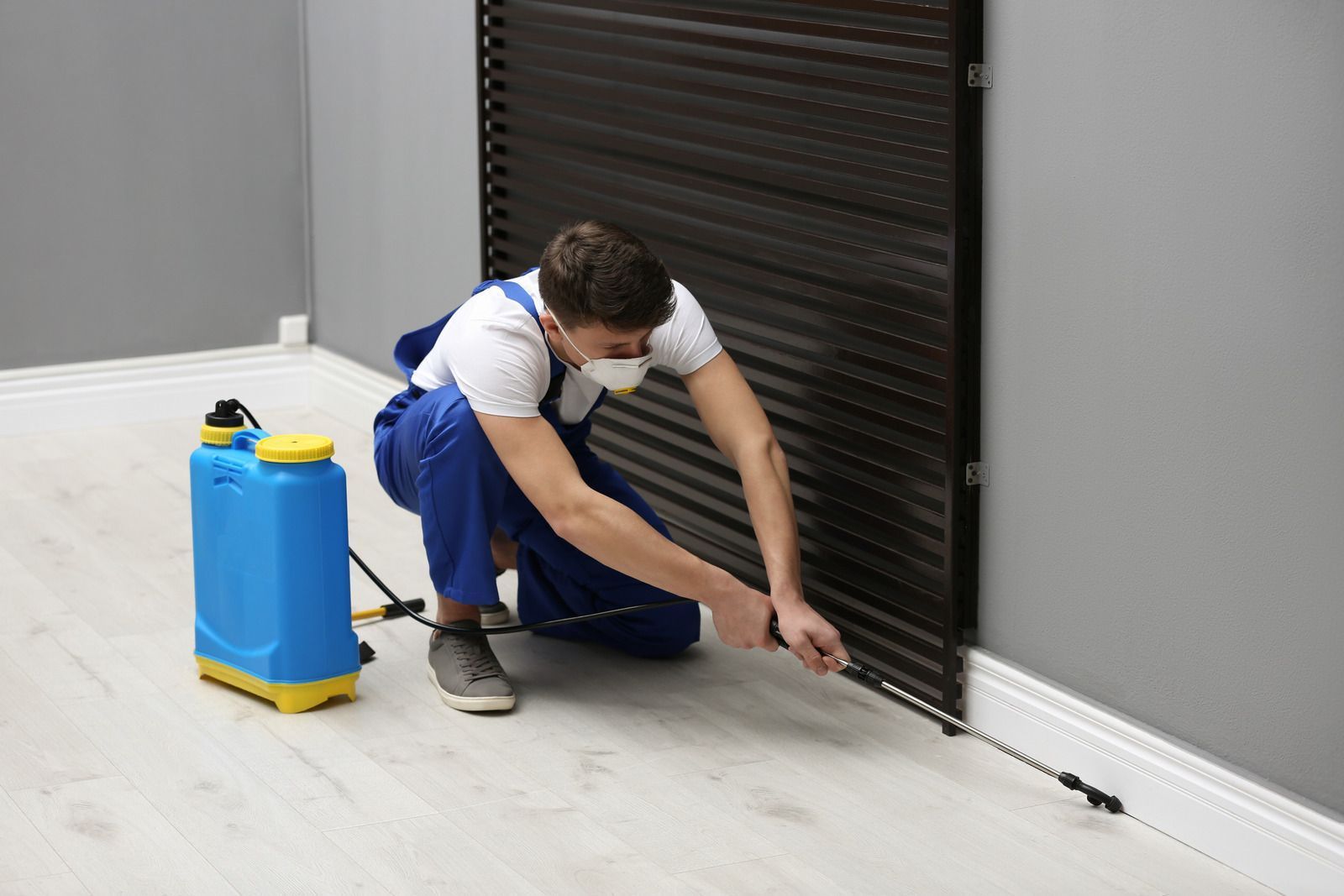 Pest control worker spraying insecticide along a wall, wearing a mask and overalls, with a blue sprayer.