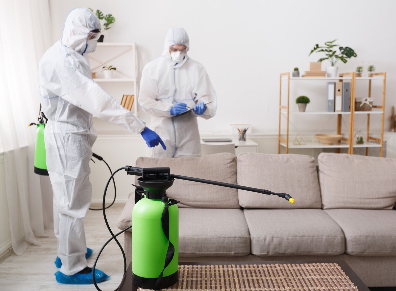 Two people in protective suits disinfect a living room with sprayers.