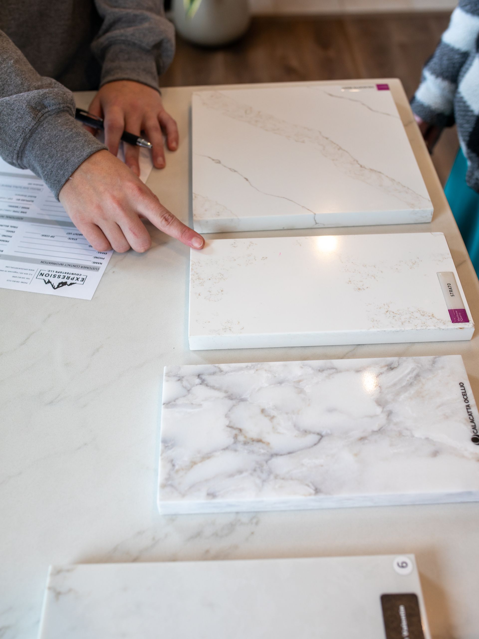 Person pointing at a white countertop sample, with other samples and a form on a white surface.