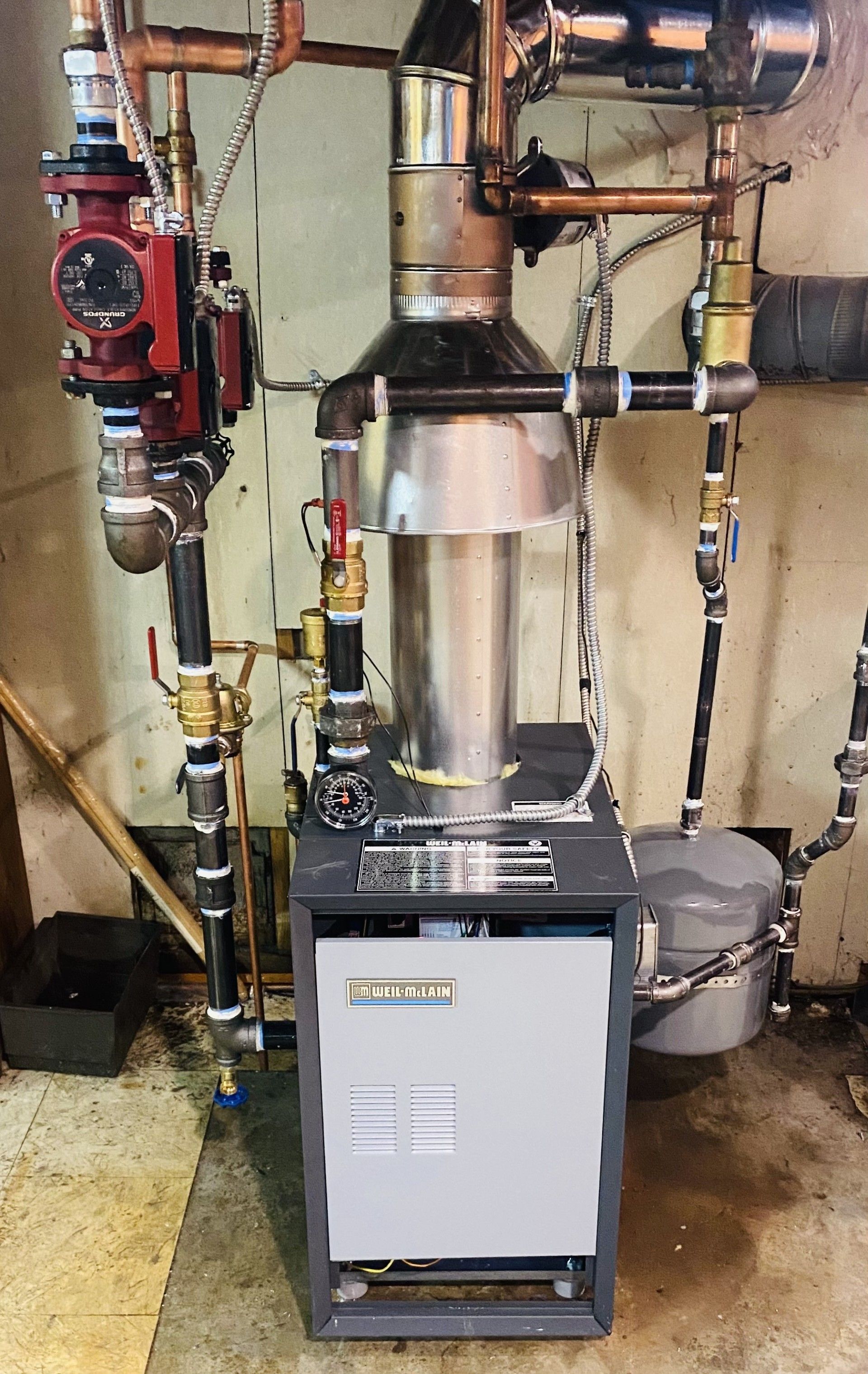 A boiler is sitting in a basement next to pipes.