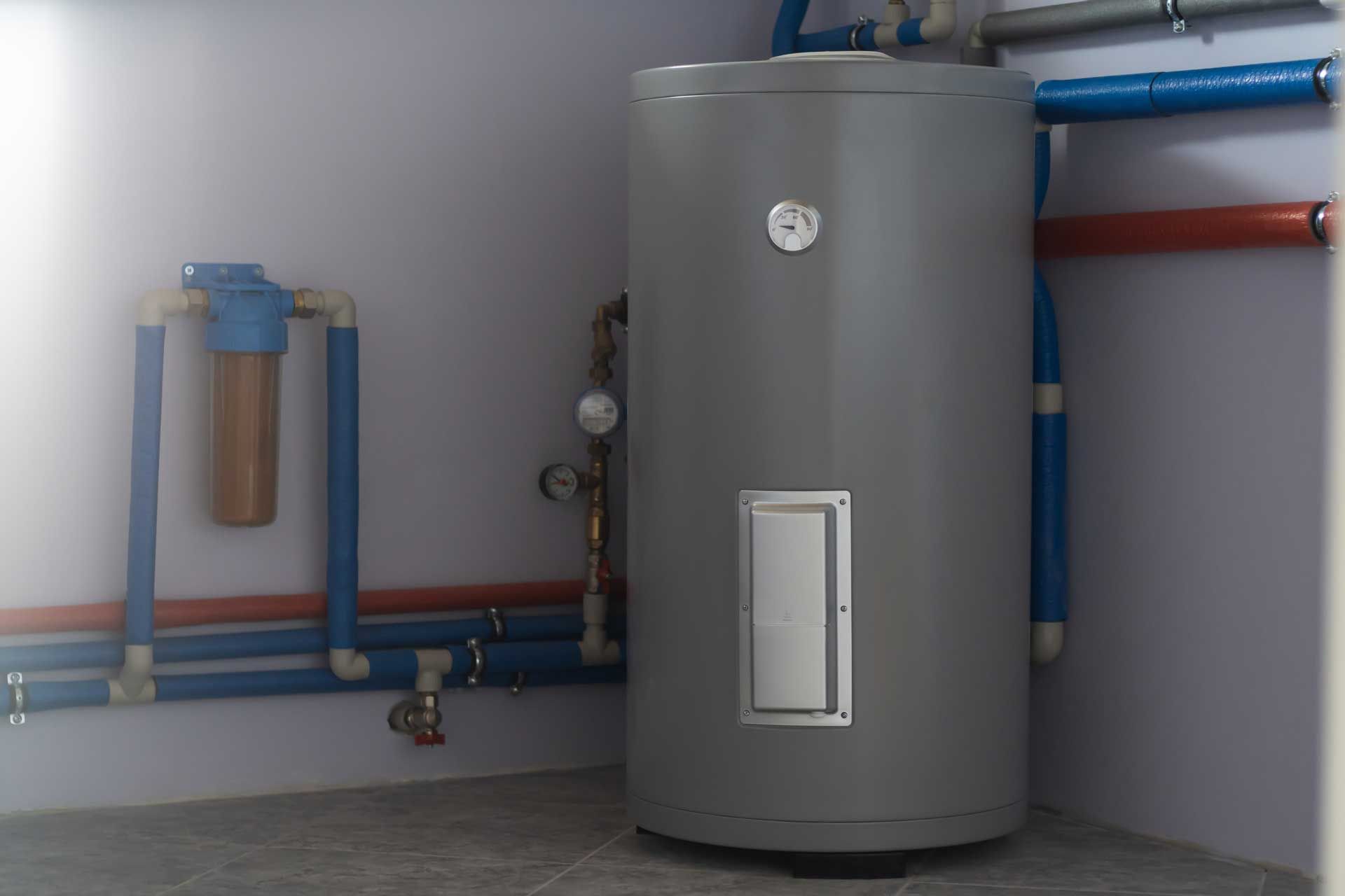 A gray water heater is sitting in a room next to pipes.
