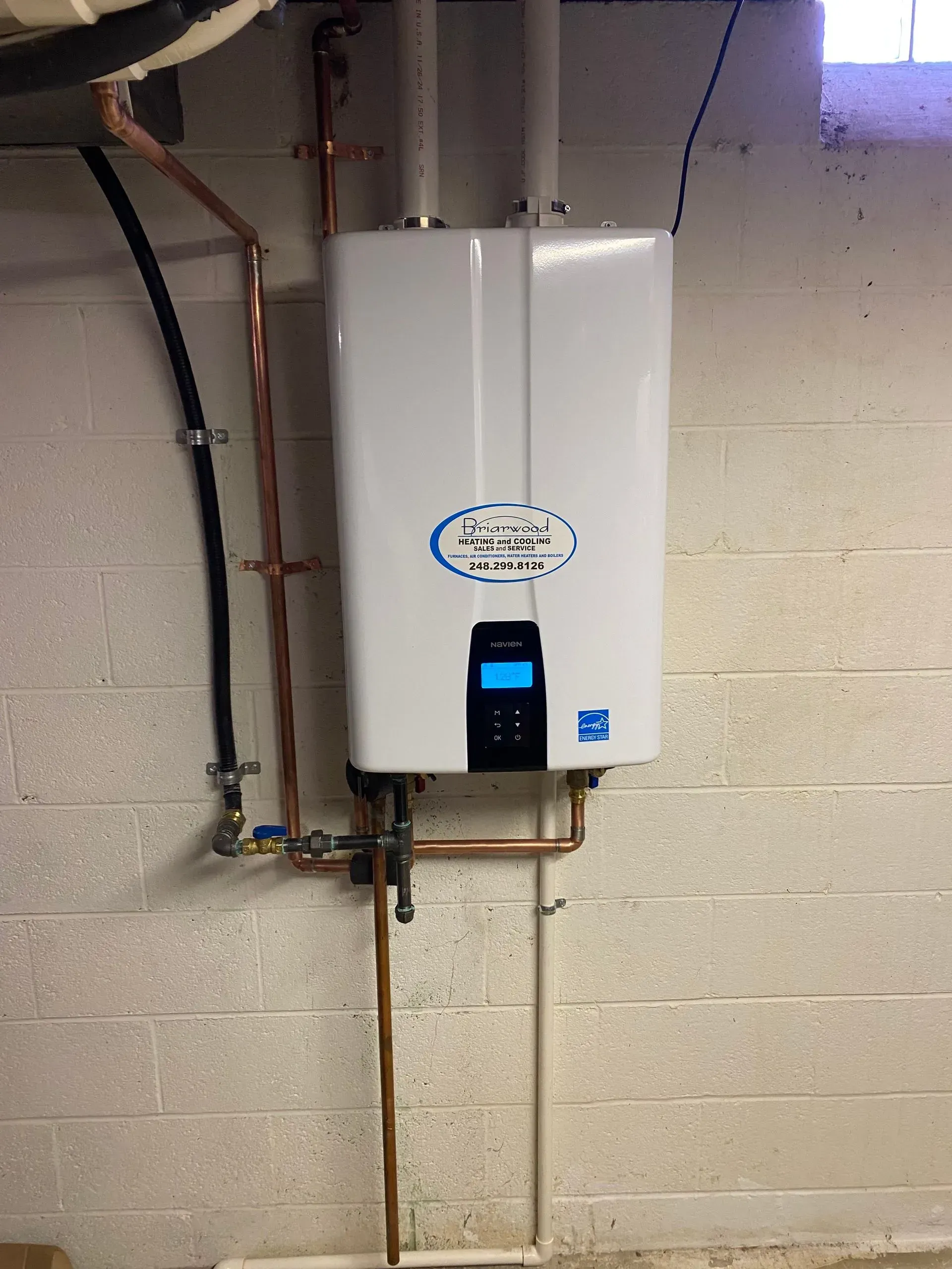A white water heater is hanging on a brick wall next to copper pipes.