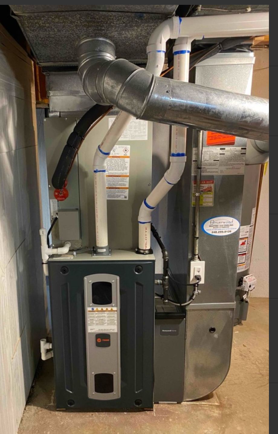 Furnace with gray, silver, and white components, including ductwork and PVC pipes, in a basement setting.