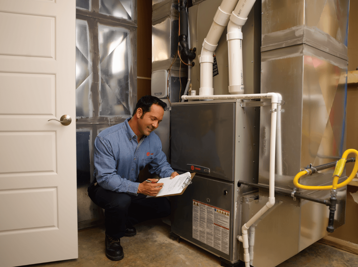 A man is kneeling down in front of a furnace in a basement.