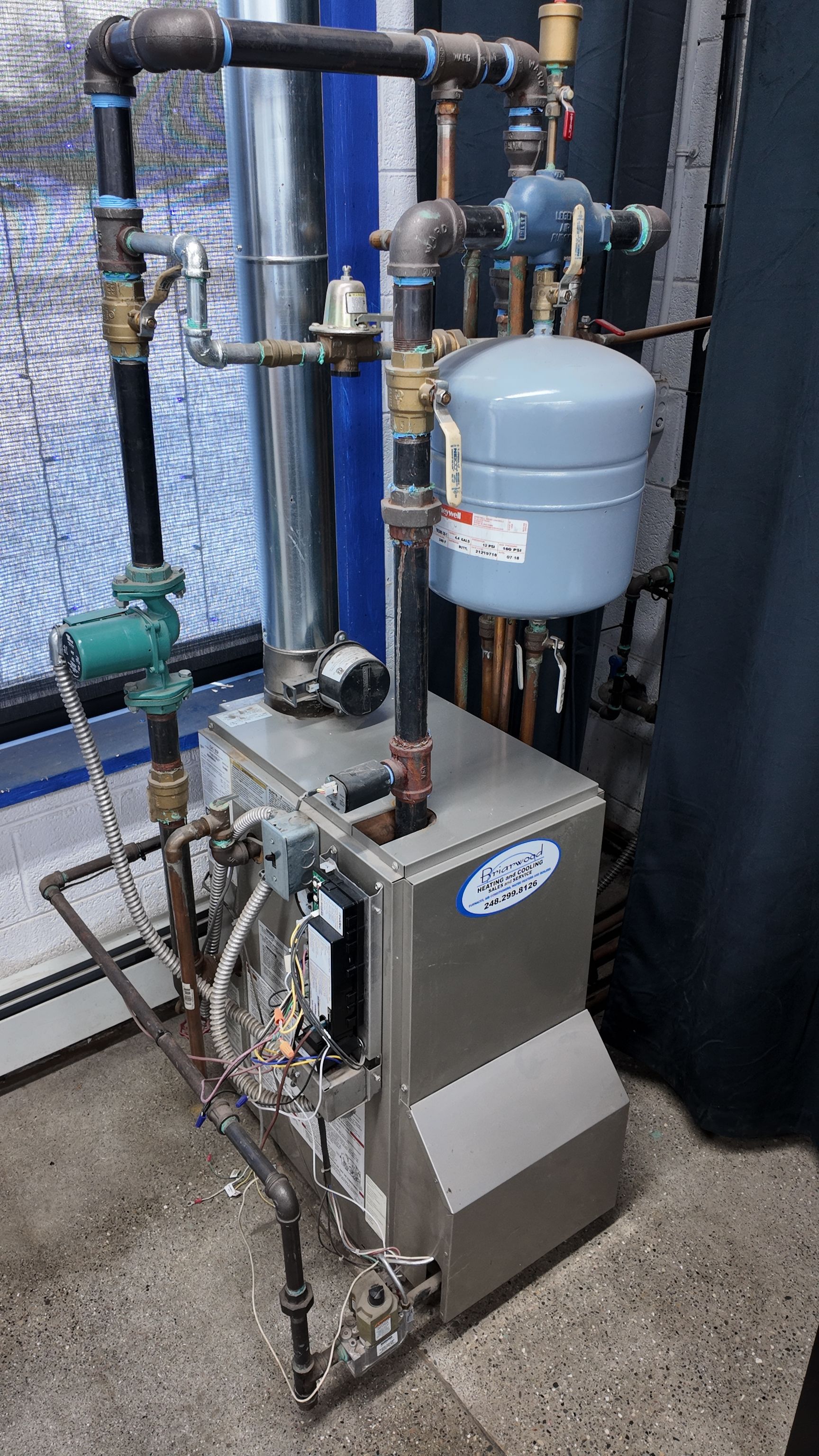 A silver HVAC boiler with pipes and a blue expansion tank against a wall.