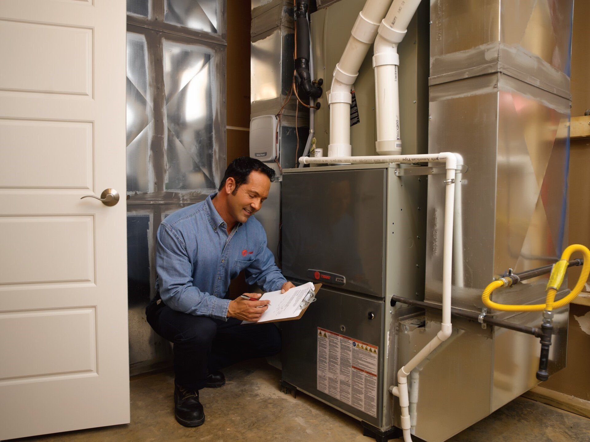 A man is kneeling down in front of a furnace and looking at a clipboard.
