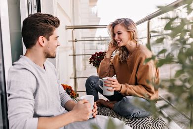 A couple is sitting on an apartment couch, holding a coffee mug, and laughing at each other.