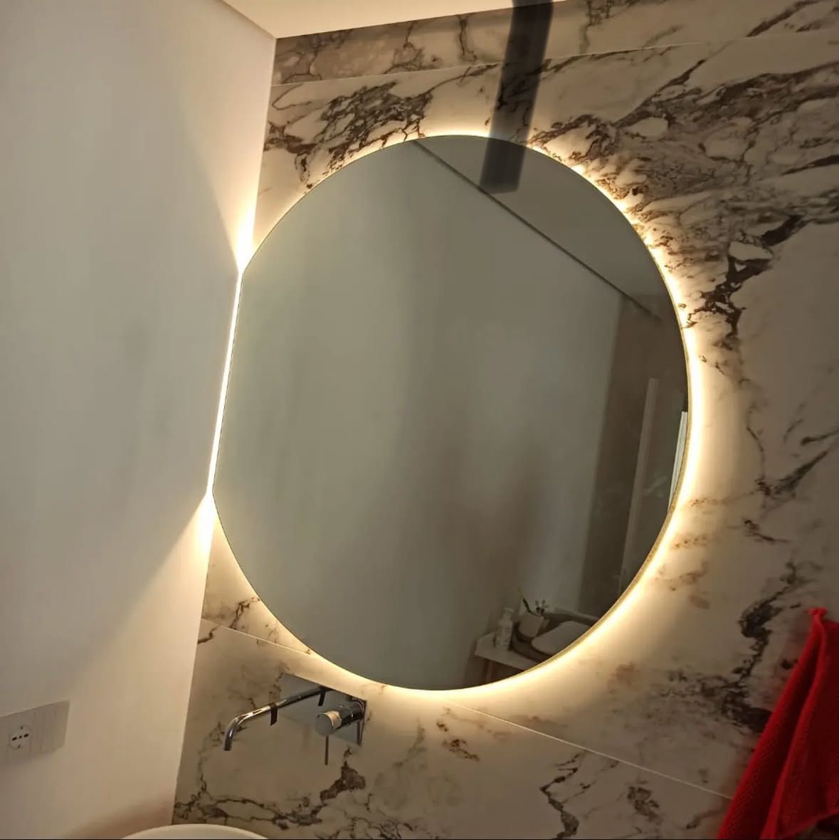 Specchio rotondo con illuminazione a LED, montato su una parete in marmo in un bagno. Sotto lo specchio c'è un rubinetto.