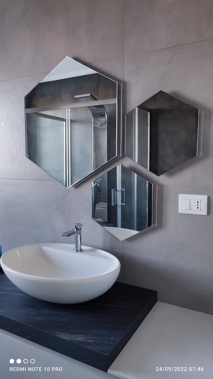 Bagno con specchi esagonali, lavandino bianco su bancone nero e pareti grigie.