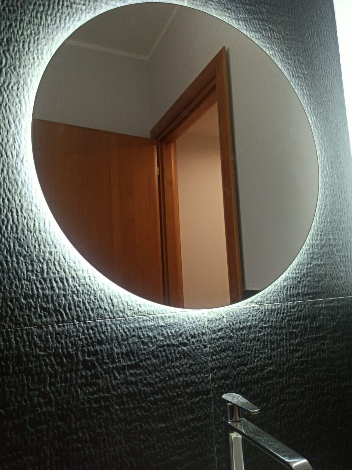 Specchio rotondo con bordo illuminato a LED, montato su una parete nera testurizzata; sotto c'è un rubinetto.