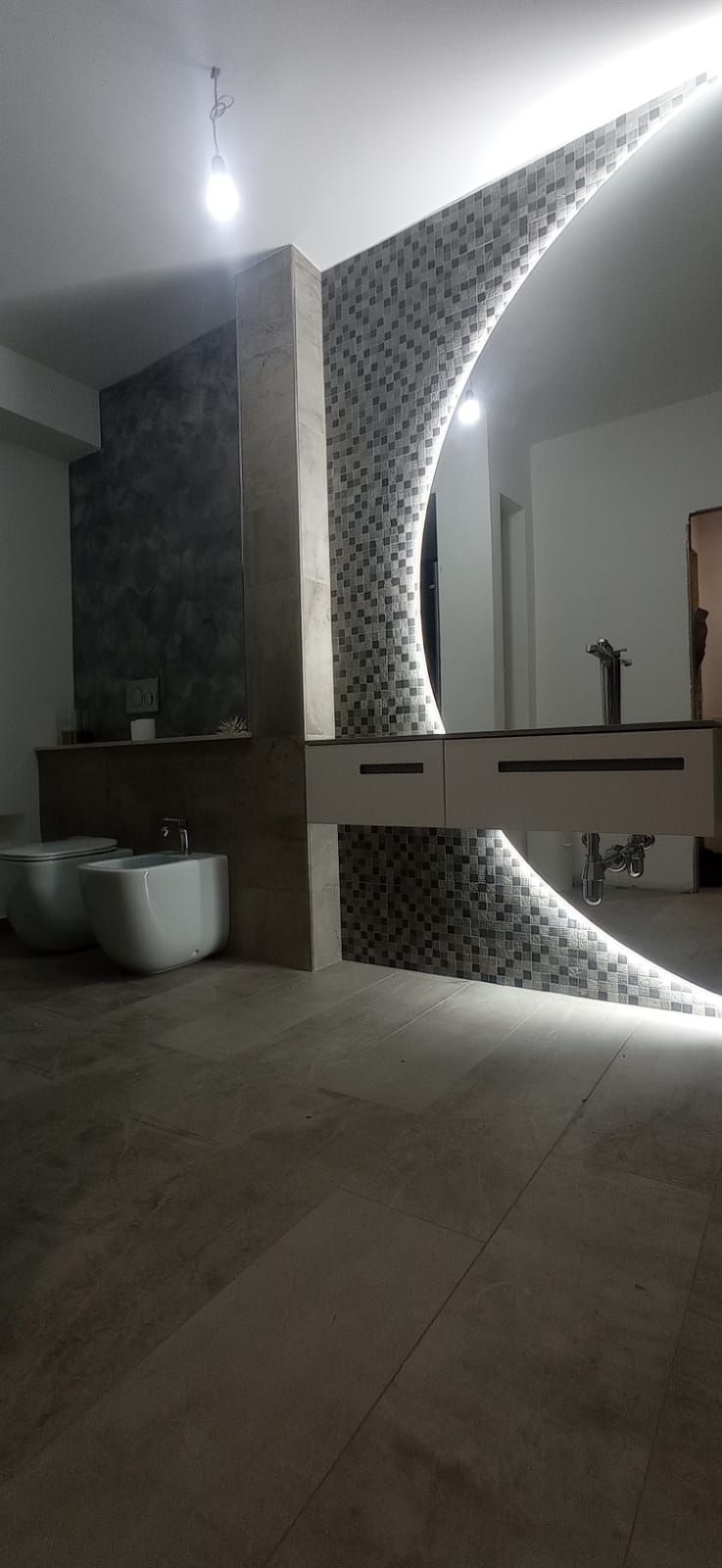 Bagno moderno con parete a mosaico, lavabo, bidet e pavimento in legno. Illuminazione intensa, design unico.