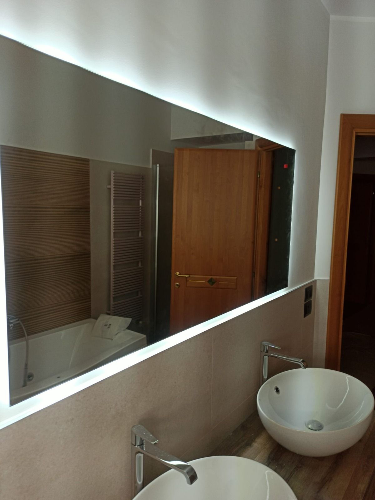 Bagno con grande specchio illuminato sopra due lavandini. Si riflettono una vasca e una porta.