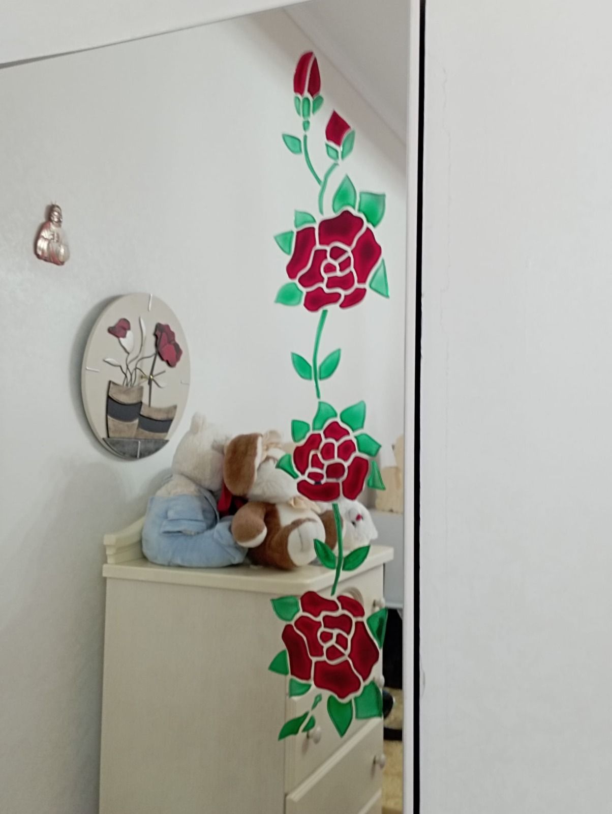 Specchio con motivo a rosa rossa, adiacente a una cassettiera bianca con un animale di peluche e un'opera d'arte sulla parete.