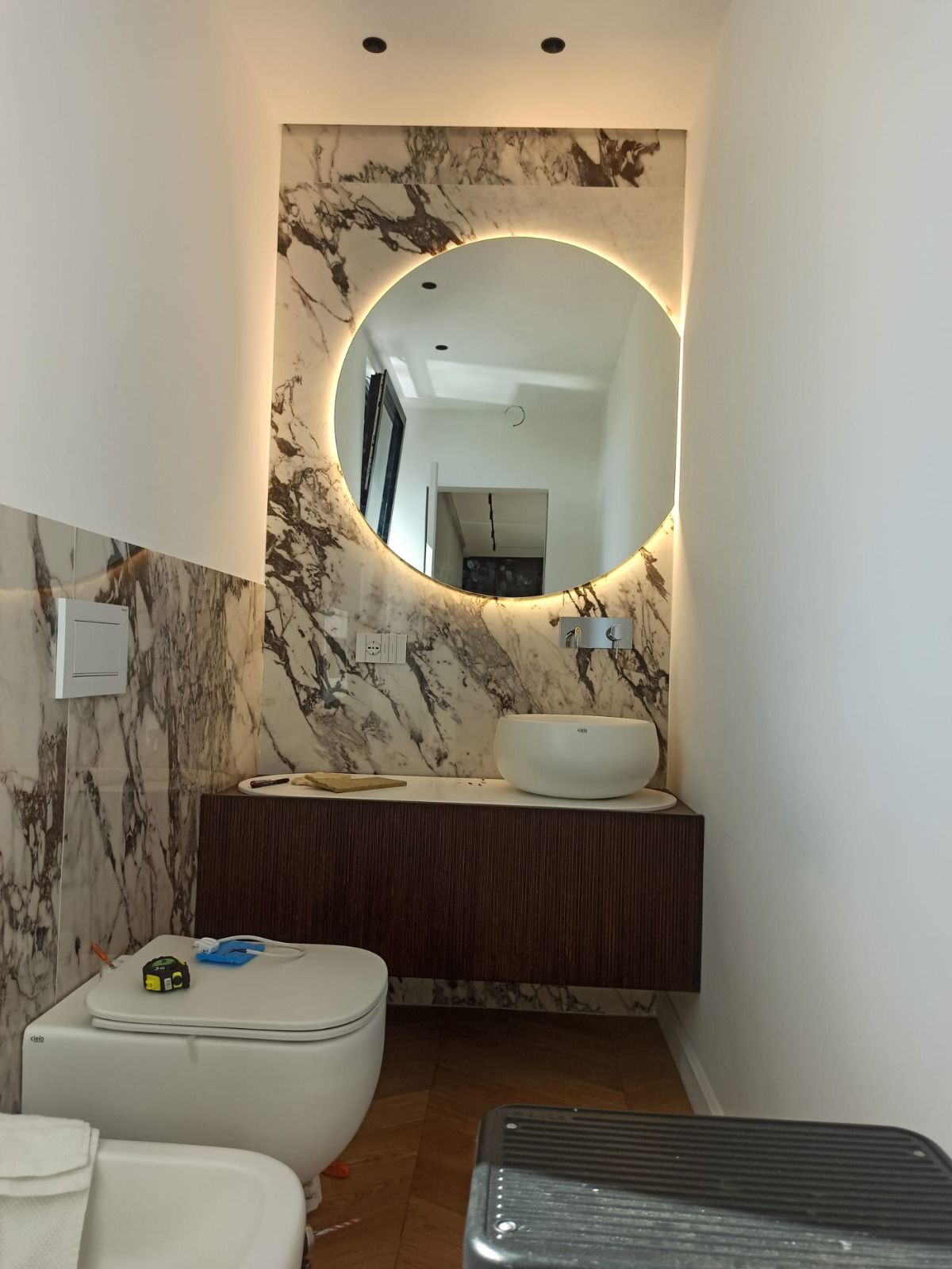 Un bagno moderno con parete in marmo, specchio circolare con illuminazione a LED, mobile lavabo in legno sospeso e WC.