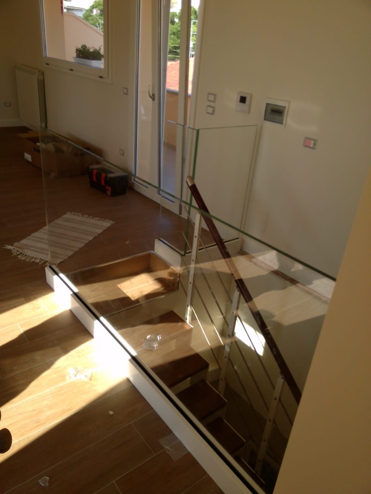 Scala con gradini in legno e ringhiera in vetro, vista dall'alto, con la luce del sole che entra a fiotti.